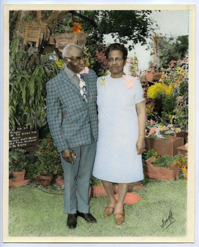 Two people in their Sunday best stand in front of a display of greenery and colorful flowers