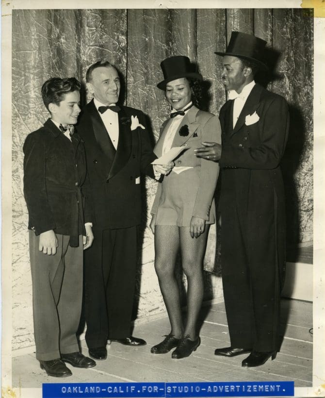 Eudora Proctor in dance outfit standing next to two men and a boy, caption: 'Oakland Calif. For studio advertisement' [ 095 ] 1949