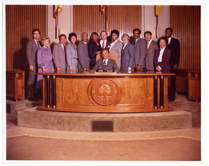 Lionel J. Wilson sits behind the mayor's desk with seceral people lined up behind him