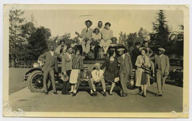 A group of well-dressed people stand around and sit on a vintage automobile