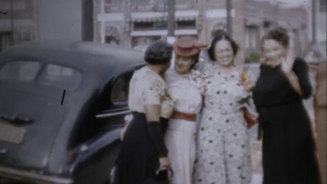 Four women stand next to an automobile in a still from a home movie