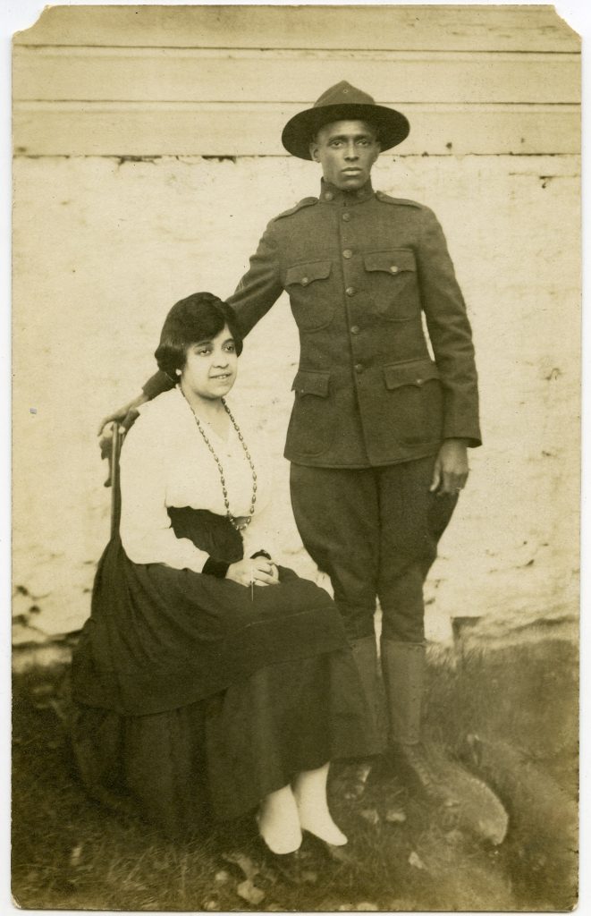 A woman is seated next to a standing man in a World War 1 - style uniform