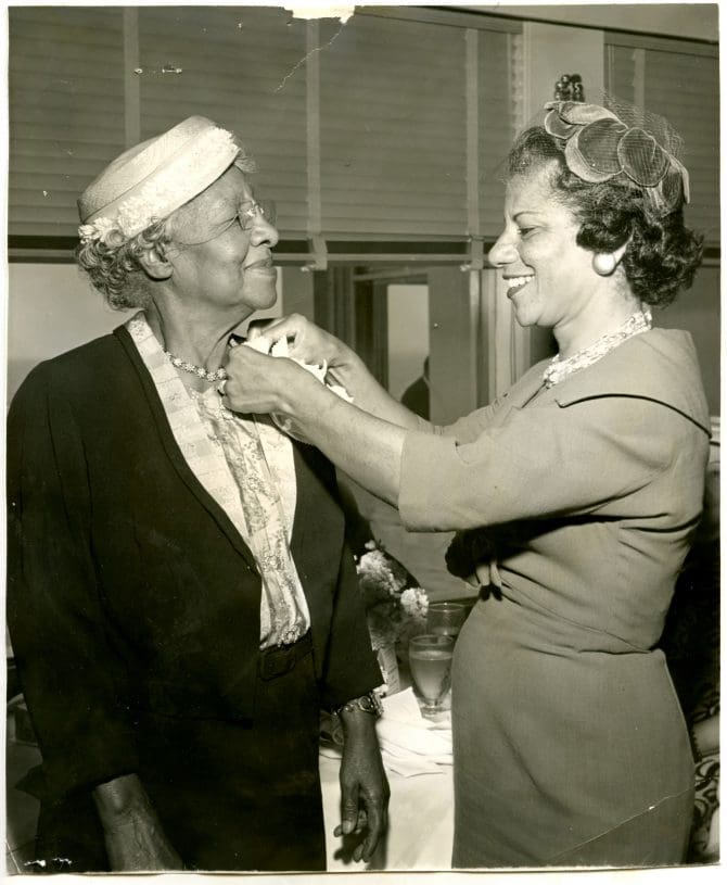 Woman pinning corsage on Sylvia Anna Magruder at her 80th birthday day breakfast given by the Society of Classical Arts at the Cliff House in San Francisco, California