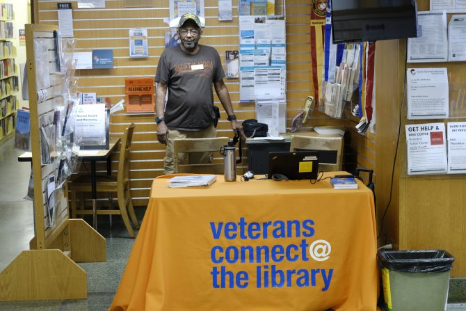 A man in a brown shirt and camoflauge hat stands behind a table with an orange tablecloth with the text "Veterans Connect @ The Library"
