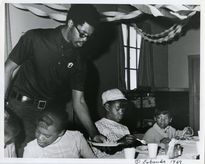 Black Panther Party member serves paper plate of food to children seated at table