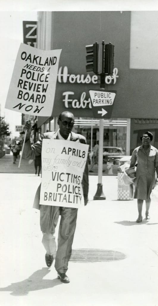 Luther Smith wearing 'on April 8 my family and I were victims of police brutality' sandwich board and waving 'Oakland needs a police review board now' sign
