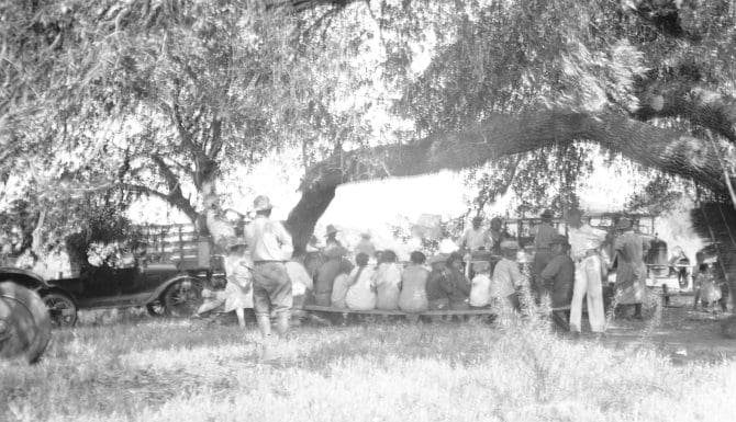 Group sitting in field, automobile parked to the left, circa 1920s