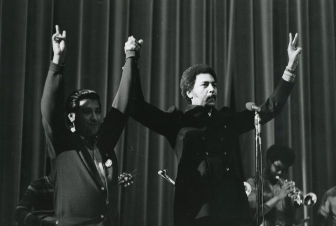 Congressman Ronald Dellums and Cesar Chavez standing on stage flashing V sign 1972-08-10