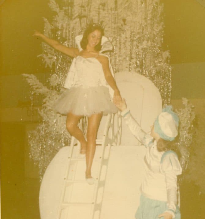 Teenager in white tutu and crown climbing down a ladder leading out of a large snowball. Another teenager in a blue and white costume, holds her hand as she descends.