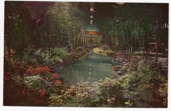 Color image of a long pool surrounded by flowers and trees, with a statue and small building in the background.
