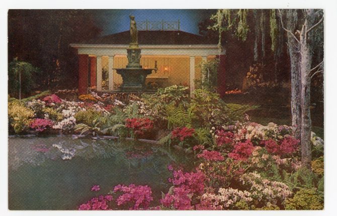 Color image of a pool surrounded by flowers and ferns, with a classically inspired statue of a woman and a small building at the edge of the pool.