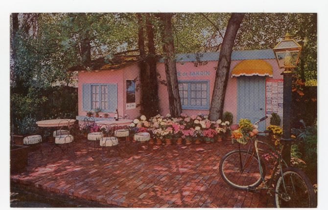 Color image showing a courtyard in front of a pink and blue cafe named Cafe de Jardin. Courtyard has several large trees surrounded with potted pink and white flowers.
