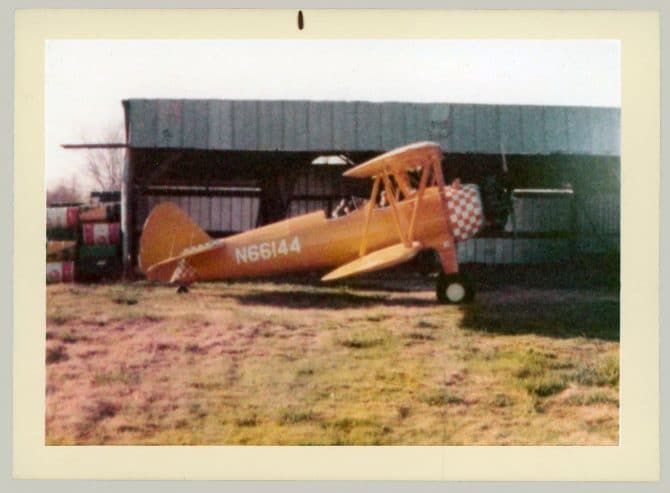 Vintage early 1970s photo of orange biplane on grass in front of metal shed