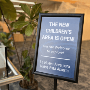 Navy blue sign with white letters stating "The New Children's Area is Open: You are Welcome to Explore" in English and Spanish, next to a fig tree, positioned at the entrance to the Children's Area