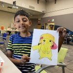A young Indian boy smiles and holds up a colored-in drawing of a Pokemon