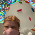 A woman wearing a mask poses for a selfie in front of a giant ice cream cone balloon art sculpture at CPL's Ice Cream Adventure celebration