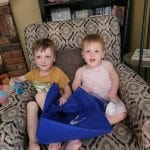 Two young children sit on a patterned armchair holding a Canton Public Library prize patrol swag bag