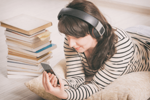 A woman wearing headphones lies on a pillow on the floor, looking at her smartphone. A pile of books sits next to her.