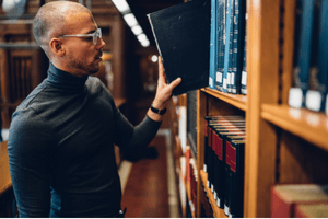 A man wearing glasses and a black turtleneck browses books on a shelf in a library