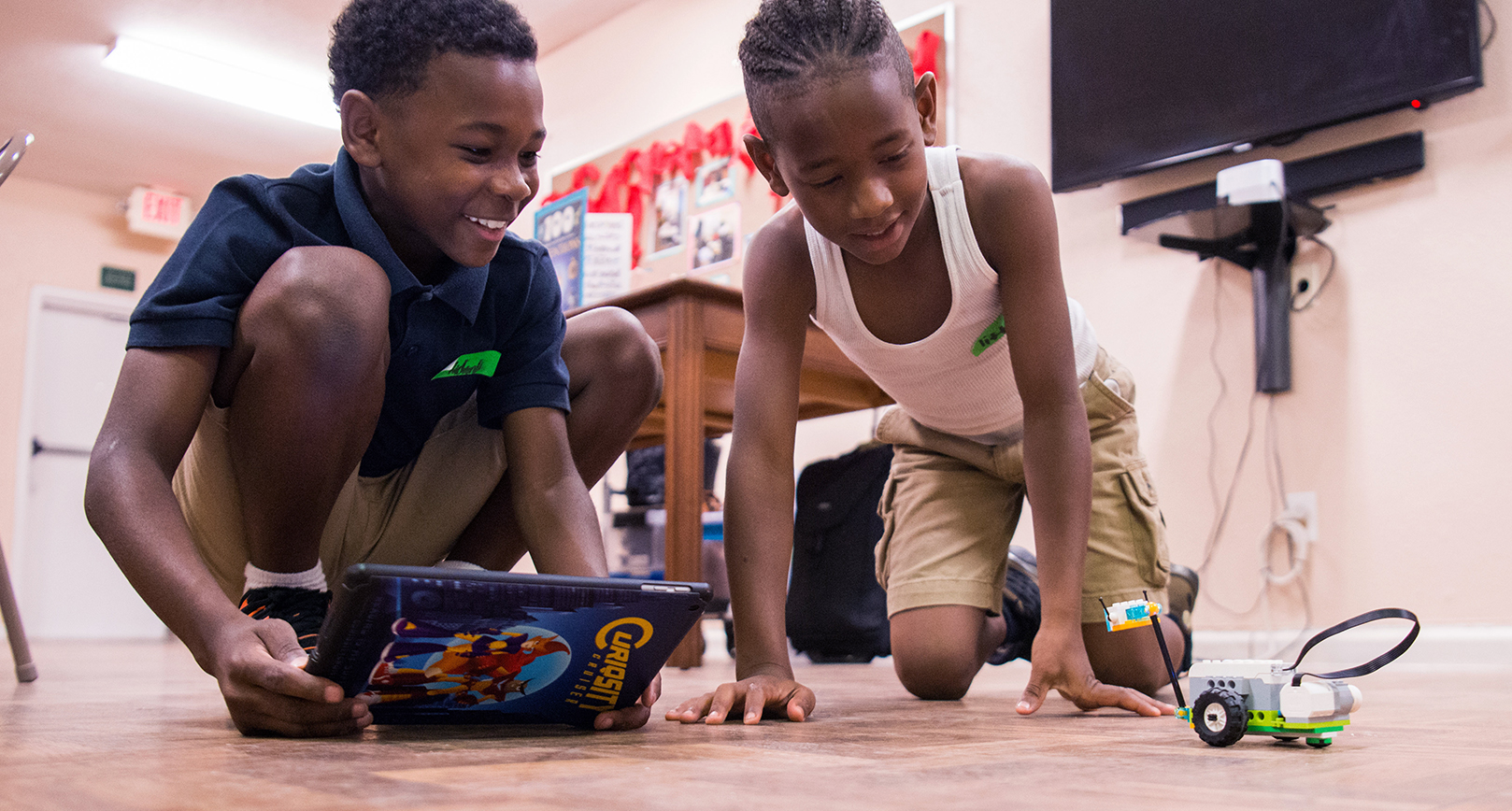 Two young boys practice coding on an iPad to control a Lego device. The children are smiling widely, sitting and squatting on the floor in a brightly lit room.