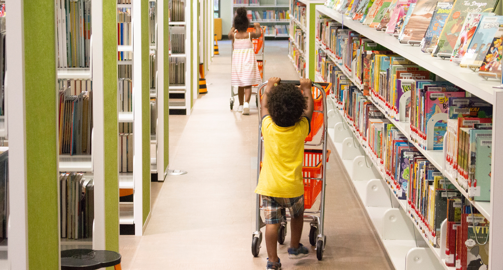 two children in the children's section of a library pushing shopping carts filled with books down the aisle