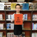 A boy holds up his "Dig Into Summer" print in front of a bookcase.