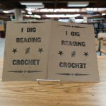 Two "I Dig Reading And Crochet" prints are displayed on a shelf, with one sign including a backwards "D" and "E."