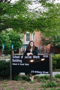 Graduate in a graduation gown smiling leaning over a sign that reads "School of Social Work Building" with tree branches reaching overhead