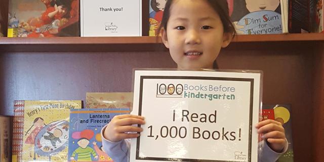 young, smiling girl holding up sign, 'I Read 1,000 Books'.
