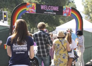 A photo taken from behind a diverse group of people walking into a rainbow-arched entry to the Silicon Valley Pride LGBTQ Festival space. In the foreground is long-haired person wearing a t-shirt with the San Jose Public Library's Summer Learning logo.