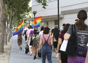 A photograph taken behind a group of people walking on a city street following two rainbow flags in the distance.