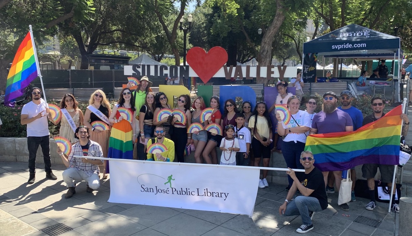 Large group of San Jose Public Library staff members smiling, celebrating, and carrying rainbow flags and fans at the Silicon Valley LGBTQIA+ Pride Festival.