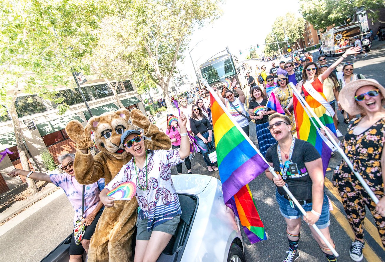 A large group of happy smiling people, including one dressed in a cougar animal suit, waving rainbow flags at a LGBTQIA+ Pride Parade.