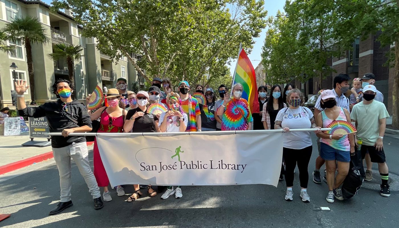 Diverse group of library staff members and their families pose for a picture at Silicon Valley Pride Parade in 2022. They are holding a white banner that reads San Jose Public Library. There is a rainbow flag in the middle with some folks holding rainbow fans.