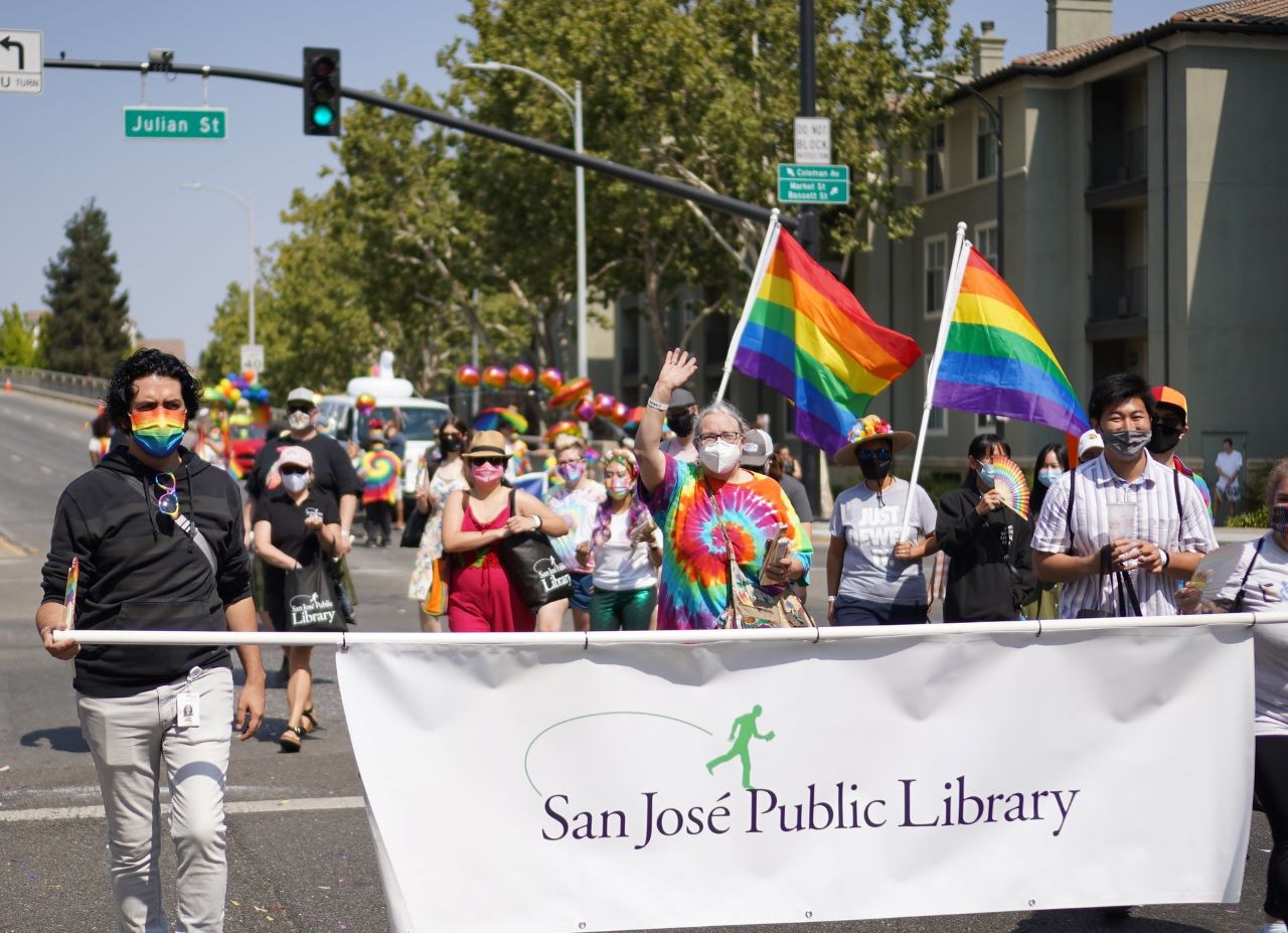 A group of San Jose Public Library staff are photographed marching in the 2021 Silicon Valley Pride Parade holding rainbow flags and a white banner that reads San Jose Public Library.