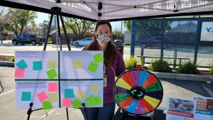 Vineland Librarian Stephanie stands at a tabling tent outside with an answer and ask board and a spinning prize wheel.