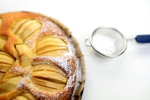 Un primer plano fotográfico de un pastel de manzana bellamente preparado, con un colador lleno de azúcar en polvo para espolvorear la parte superior del pastel de manzana.