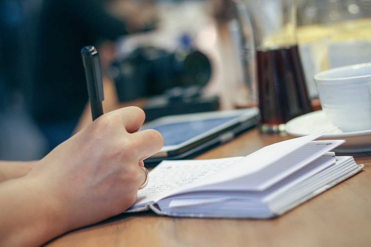 Person writing in a notebook with coffee cup and a laptop in the background.
