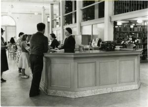 The circulation desk of the main San Jose Public Library in the 1950s. Several patrons wait in line to checkout materials. Two library staff members assist those waiting in line. On the left, Gertrude Jansens; on the right, Kathyrn Williams.