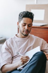 Pensive man with medium-brown skin and short dark brown hair with a short beard writing in notebook