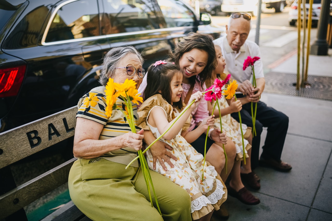 A smiling Asian family of a mother, two children, and two grandparents sitting on a city bench holding flowers.
