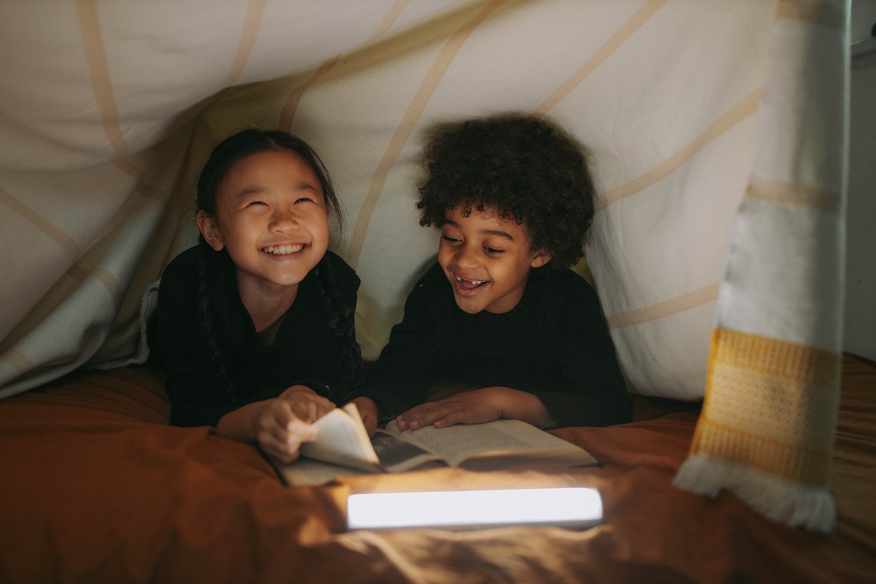 Photo of a young boy and a girl smiling while reading a book together underneath a blanket.