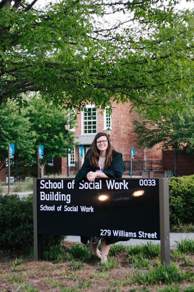 曇った日に木の下にいる、長い茶色の髪をした人が、「School of Social Work Building」と書かれた看板の上で腕を組んで微笑んでいる。