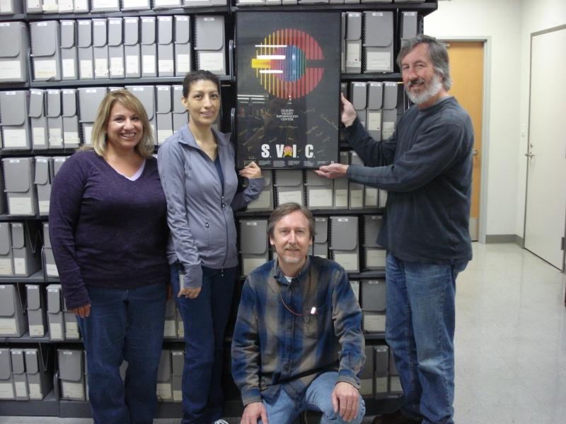SVIC で働いていた 2015 年のキング図書館スタッフ (左から) ローリー オーク、マリア ピネダ、ラルフ ピアース、リン ハリスが、SVIC コレクションと額入りの SVIC ポスターとともにポーズをとっています。