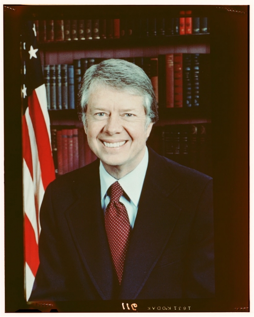 Jimmy Carter, head-and-shoulders portrait, facing front, next to an American flag., 1977.
