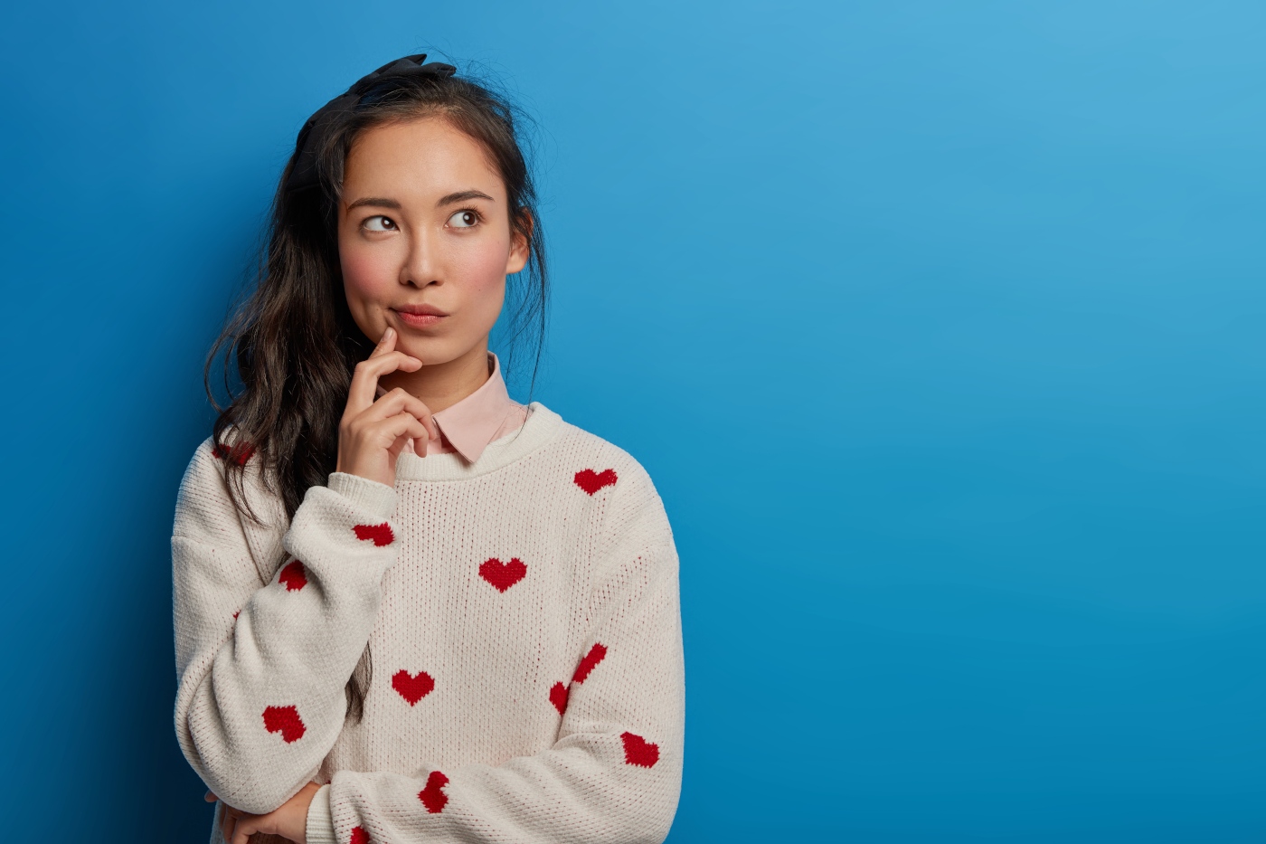 Contemplative pretty millennial young Asian woman looks away, builds plans in mind, has thoughtful face expression, long dark pony tail, wears sweater with hearts, isolated over blue background.