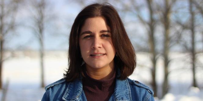 Katherine Abbass, with shoulder-length brown hair, wearing a deep maroon sweater and jean jacket, stands in front of snow and bare trees.
