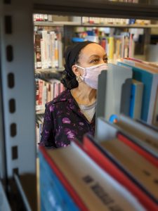 Library staff member browsing books in the library. The staff member is wearing a face mask that covers their nose and mouth.