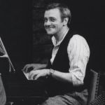 a man in a vest plays the piano and smiles