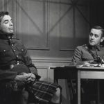 a man wearing a kilt and military jacket sits in a chair. another man in uniform, seated at a table looks at him.